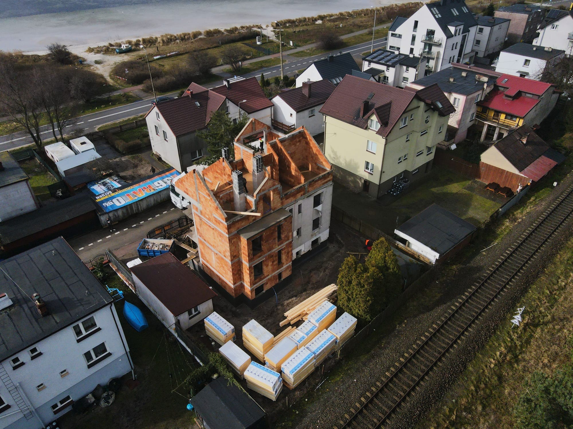 Budowa domu z czerwonej cegły na działce z widokiem z lotu ptaka. W tle morze i zabudowa miejska. Częściowo otynkowane ściany i widoczne pustaki.