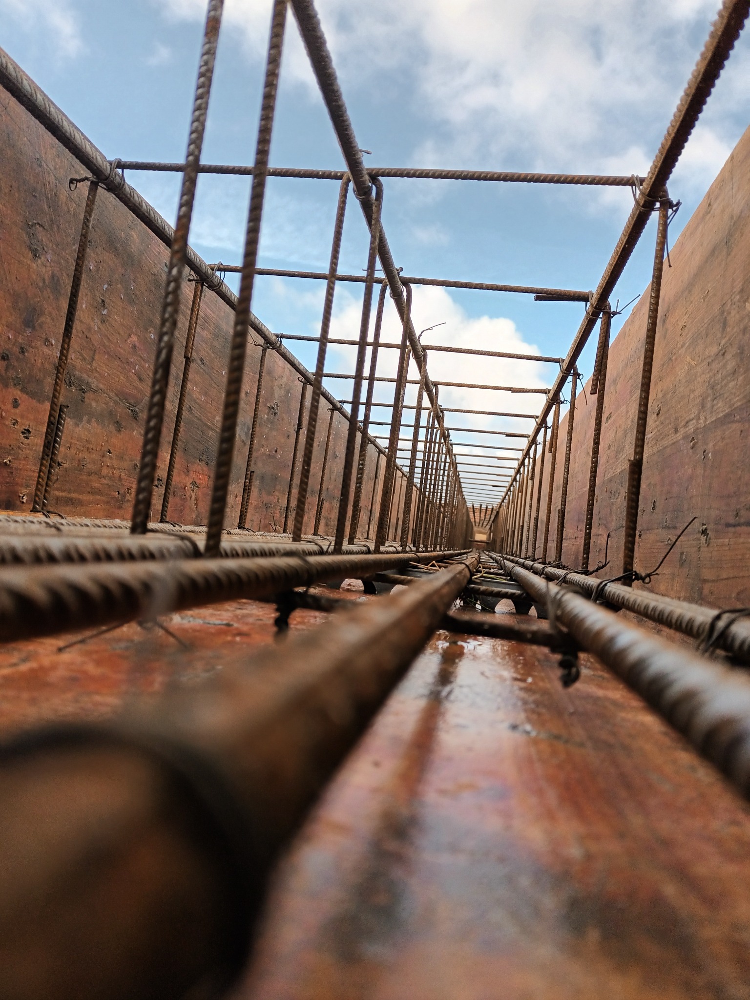 Z bliska: Zbrojenie fundamentów w drewnianym szalunku, widok w górę na niebo. Stalowe pręty tworzą siatkę wewnątrz formy, gotowe do zalania betonem.