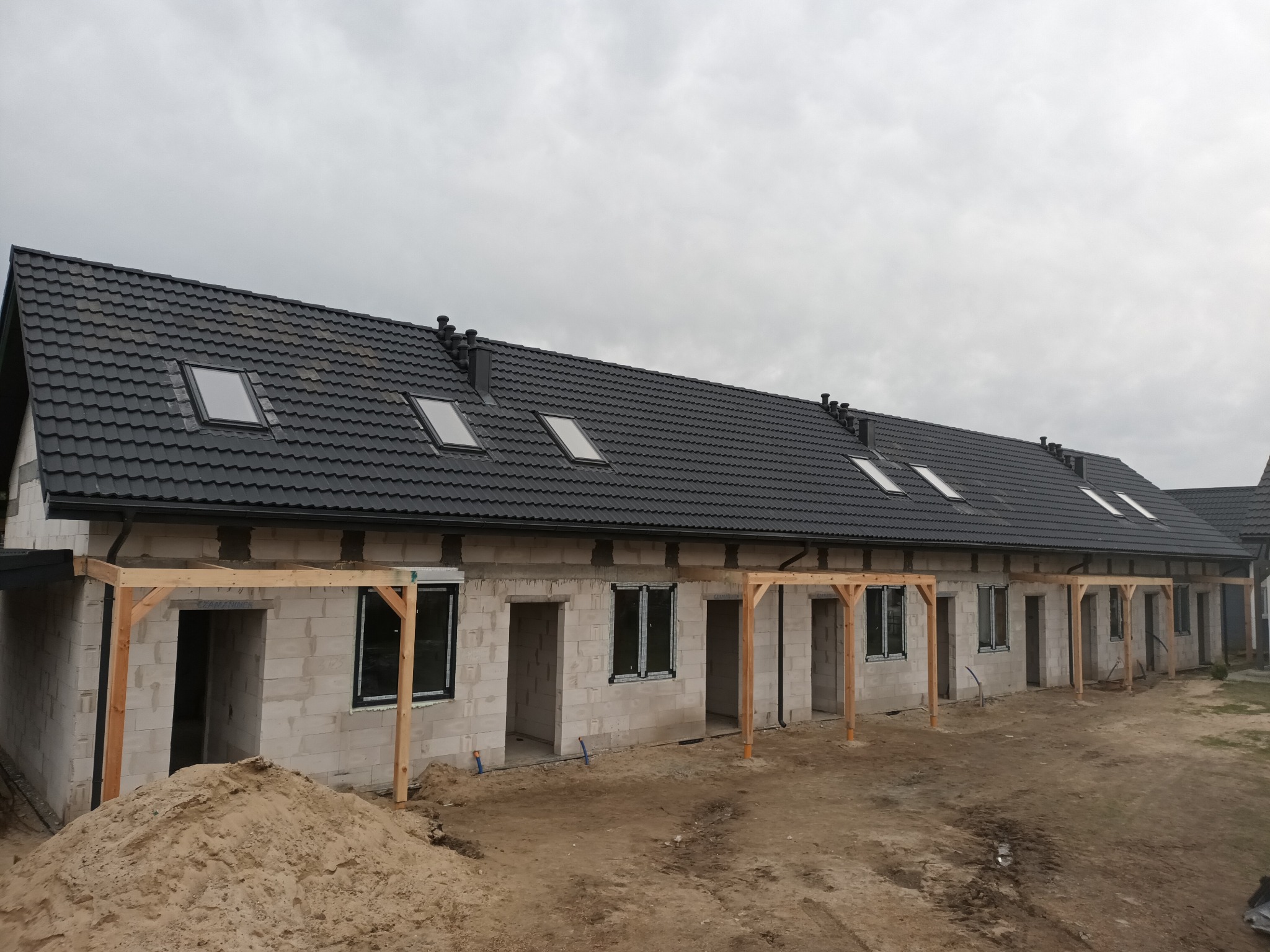 Szeregówka w stanie surowym otwartym, z oknami, dachem z lukarnami i drewnianą konstrukcją werandy. Na pierwszym planie hałda piasku.