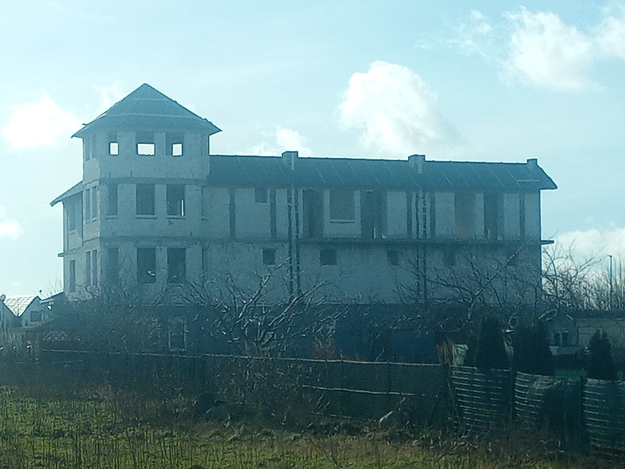 Niedokończony, trzypiętrowy budynek z wieżą w stanie surowym, widoczny zza ogrodzenia i roślinności. Brak okien, surowy beton. Błękitne niebo w tle.