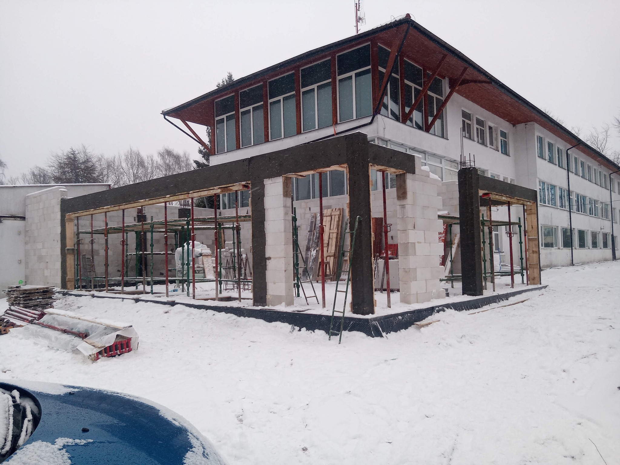Budowa domu w zimowej scenerii: wznoszone ściany z białych bloczków, widoczne rusztowania i szalunki, tło stanowi biały budynek i zasypane śniegiem otoczenie.