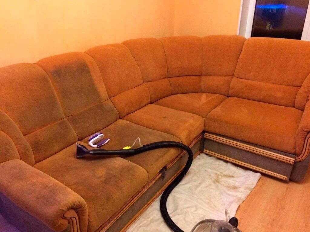 Narożnik tapicerowany w odcieniach brązu z widocznymi plamami, częściowo czyszczony odkurzaczem piorącym, na podłodze biały ręcznik z zabrudzeniami.