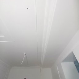 Dominik Hryciuk - Biały sufit z dekoracyjnymi listwami i wystającym kablem, widok z dołu, fragment osoby w prawym dolnym rogu kadru. Wykończenie wnętrz.