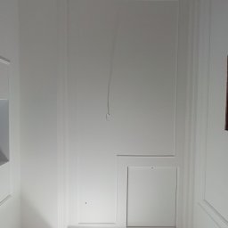 Dominik Hryciuk - Biała ściana z dekoracyjnymi listwami i dwoma włazami rewizyjnymi w Siedlcach. Widoczny przewód wystający ze ściany. Wykończenie wnętrz.