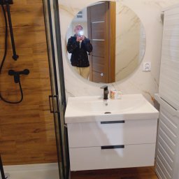 Dominik Hryciuk - Nowoczesna łazienka z kabiną prysznicową, okrągłym lustrem i białą szafką pod umywalką. Na ścianach płytki imitujące marmur i drewno. W lustrze odbicie osoby robiącej zdjęcie.