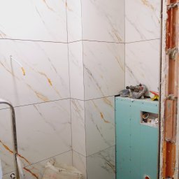 Dominik Hryciuk - Białe płytki imitujące marmur z pomarańczowymi żyłkami w łazience, częściowo wykończone. Widoczne narzędzia i płyta karton gips na ścianie.
