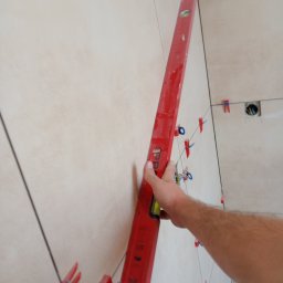 Dominik Hryciuk - Układanie jasnych płytek na ścianie z użyciem poziomicy i klipsów dystansowych. Widoczny fragment dłoni przytrzymującej narzędzie.