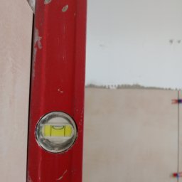 Dominik Hryciuk - Czerwona poziomica przy beżowych płytkach w trakcie układania. Widoczne krzyżyki dystansowe. Prace glazurnicze w Siedlcach.