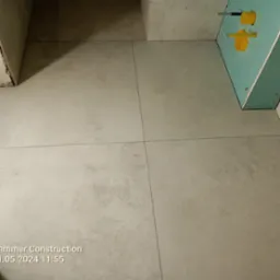 Ułożone, beżowe płytki podłogowe w łazience, widoczne fragmenty ściany z instalacją wodną. Minimalistyczny design, neutralna kolorystyka.