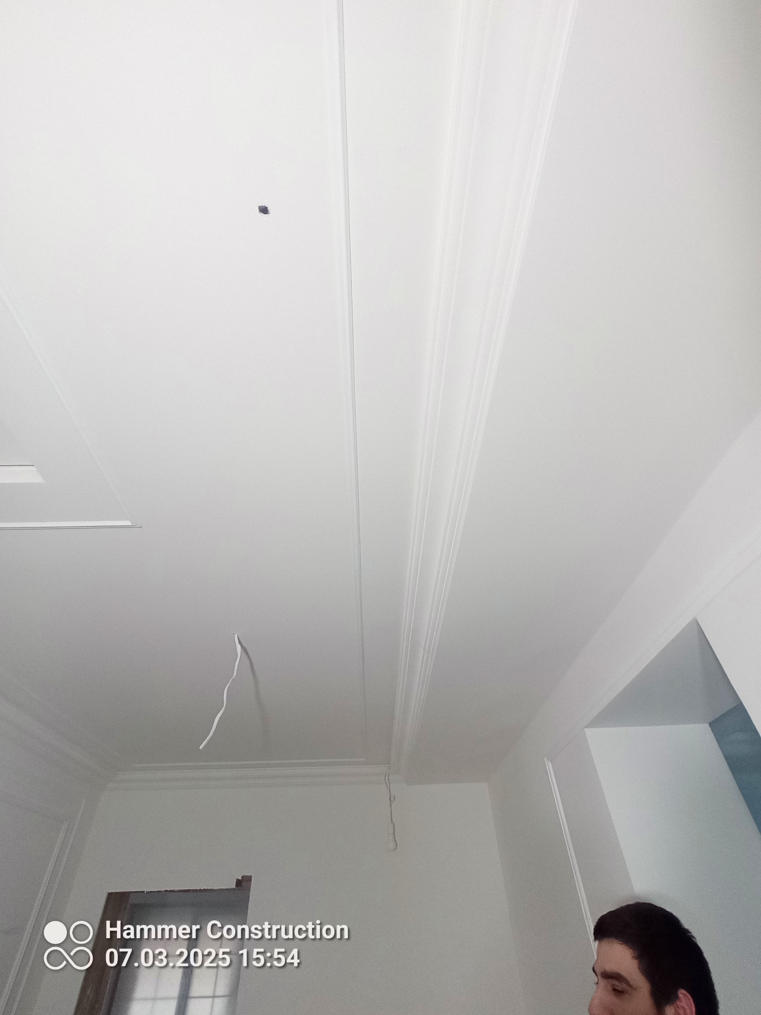 Biały sufit z dekoracyjnymi listwami i wystającym kablem, widok z dołu, fragment osoby w prawym dolnym rogu kadru. Wykończenie wnętrz.