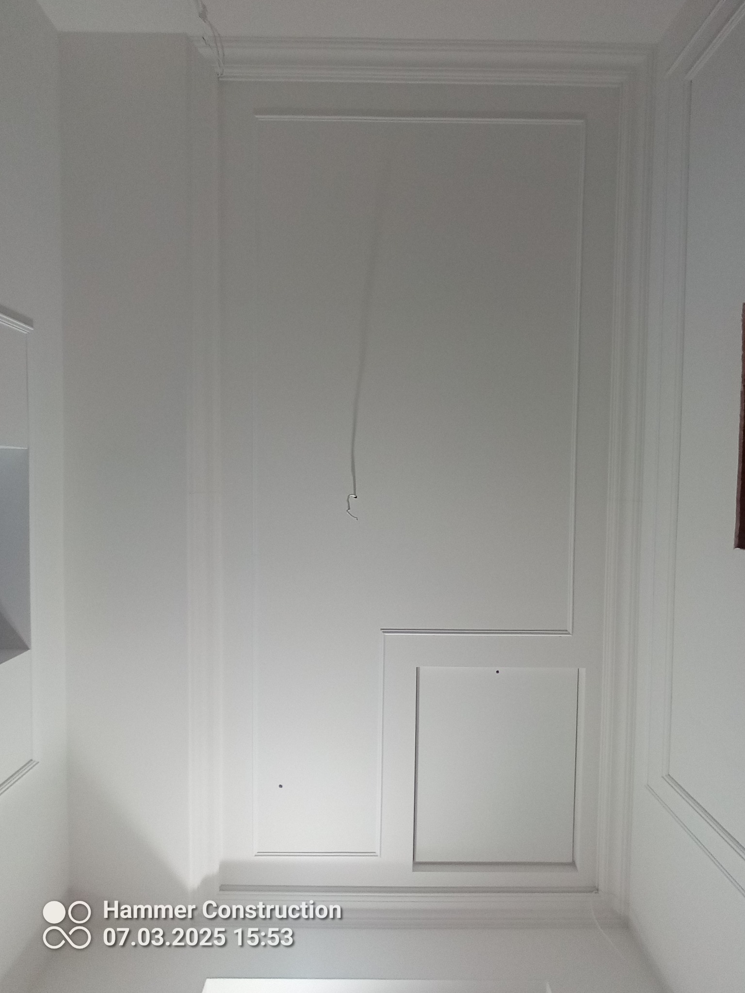 Biała ściana z dekoracyjnymi listwami i dwoma włazami rewizyjnymi w Siedlcach. Widoczny przewód wystający ze ściany. Wykończenie wnętrz.