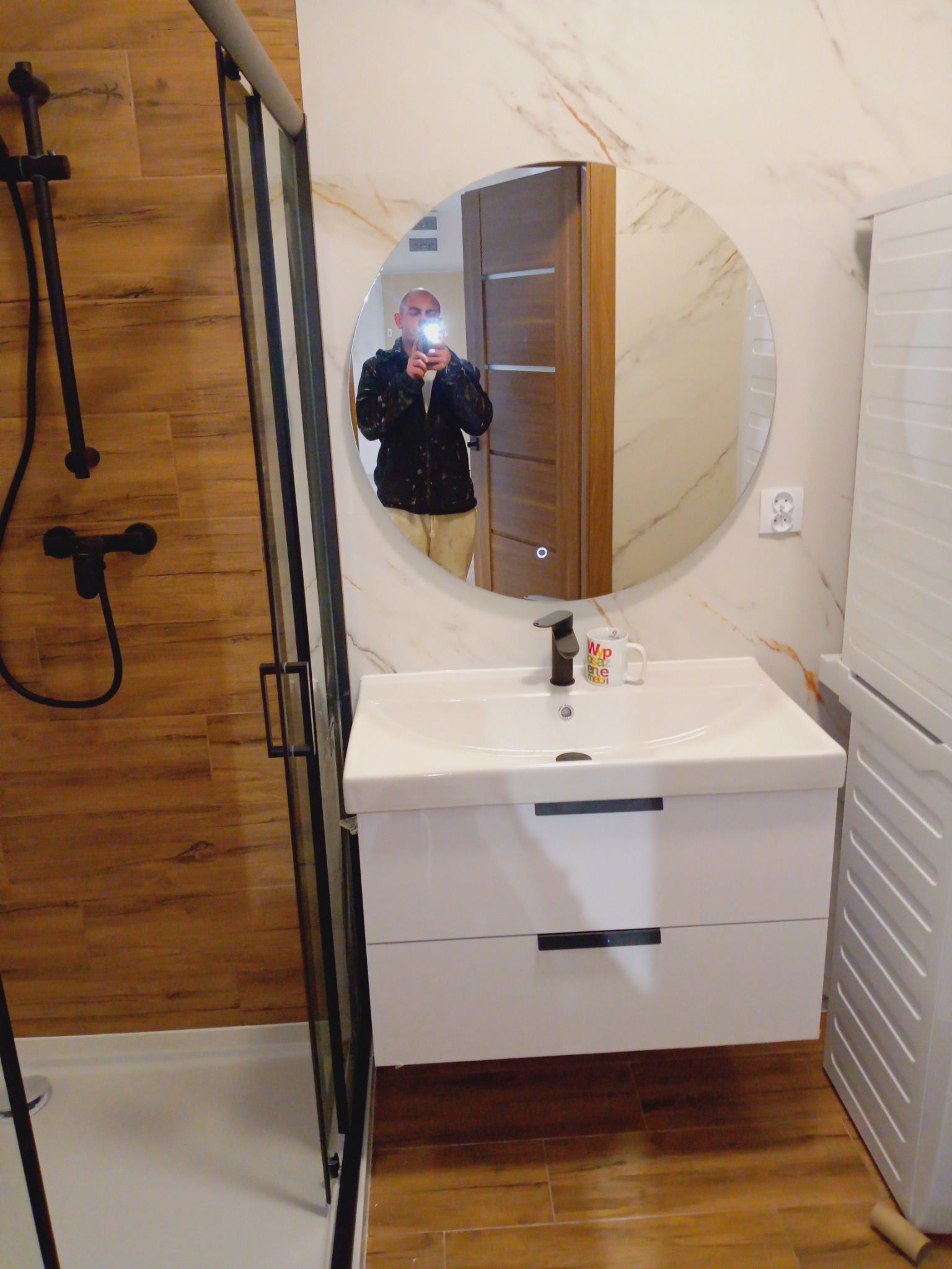 Nowoczesna łazienka z kabiną prysznicową, okrągłym lustrem i białą szafką pod umywalką. Na ścianach płytki imitujące marmur i drewno. W lustrze odbicie osoby robiącej zdjęcie.