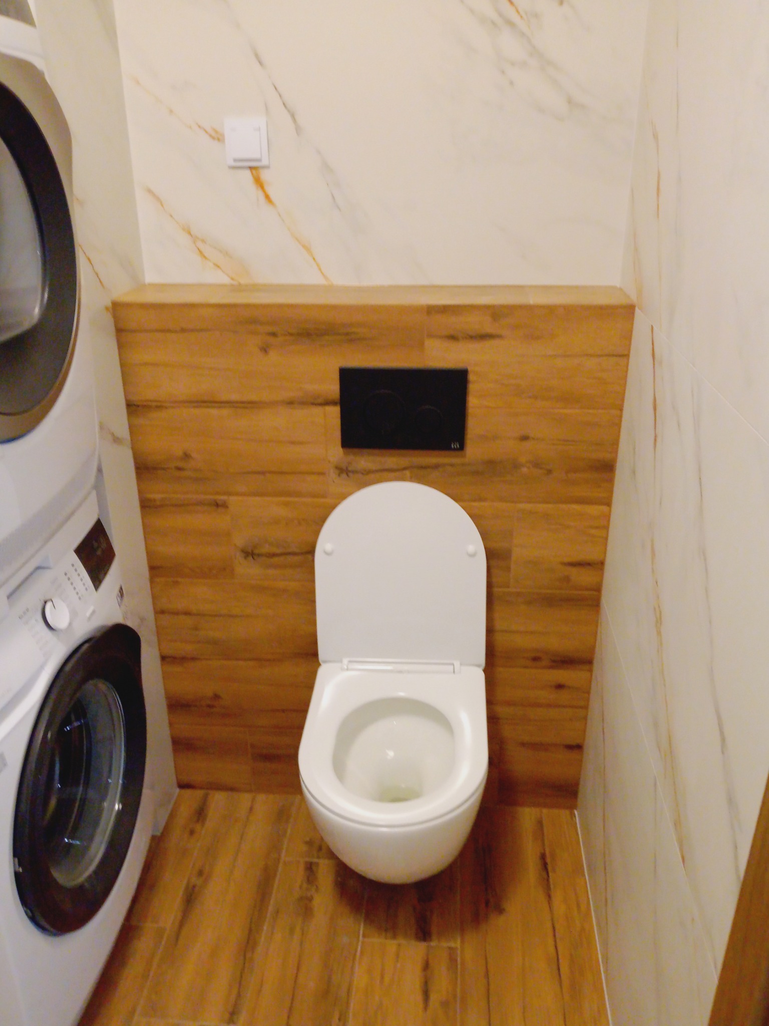 Nowoczesna toaleta podwieszana z drewnianą obudową i marmurowymi płytkami w łazience z pralką. Minimalistyczny design, ciepłe oświetlenie.