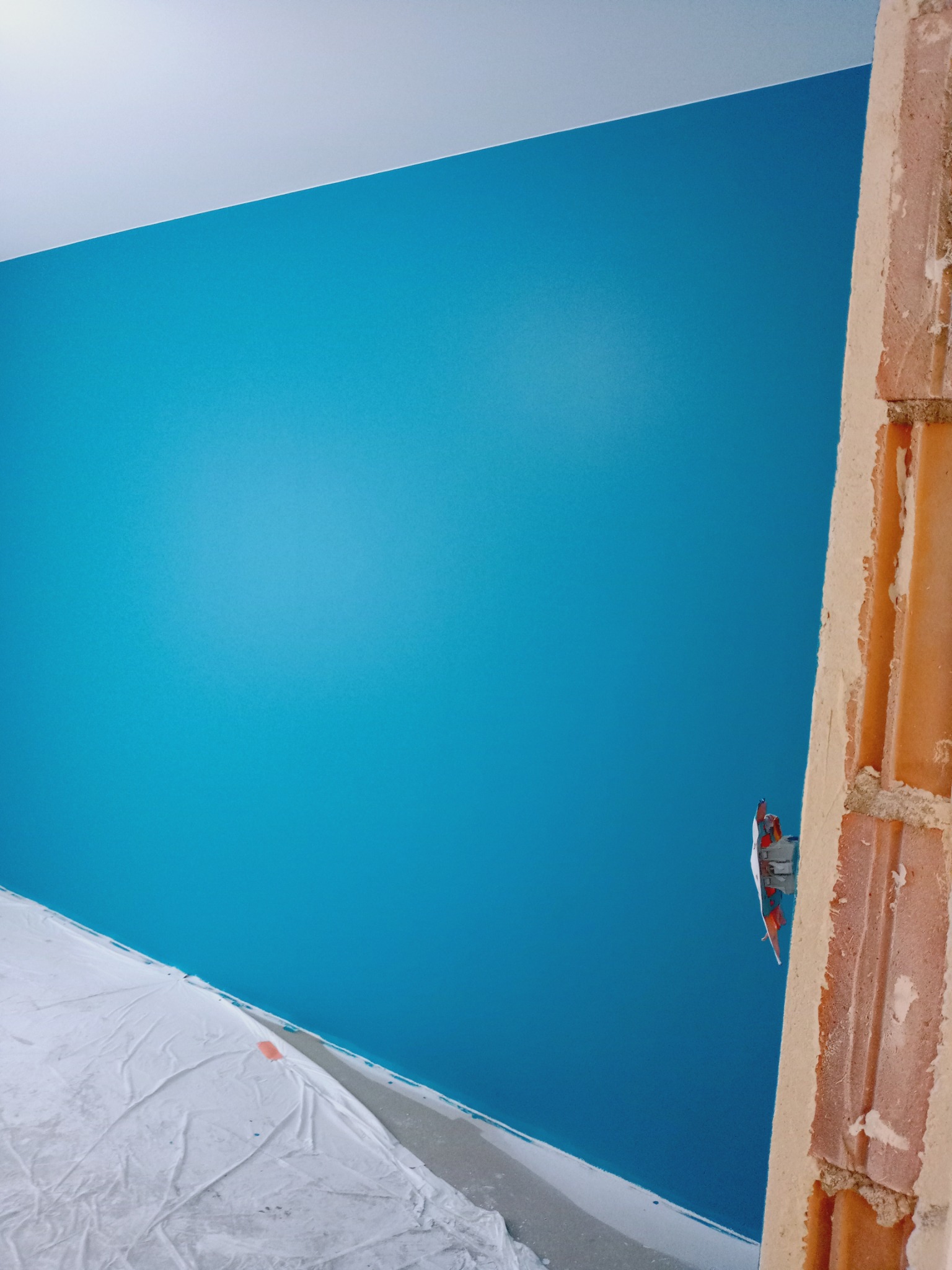 Ściana pomalowana na turkusowo, widok z dołu, z fragmentem zakrytej folią podłogi i surowym murem. Widoczna puszka elektryczna. Świeżo po malowaniu.