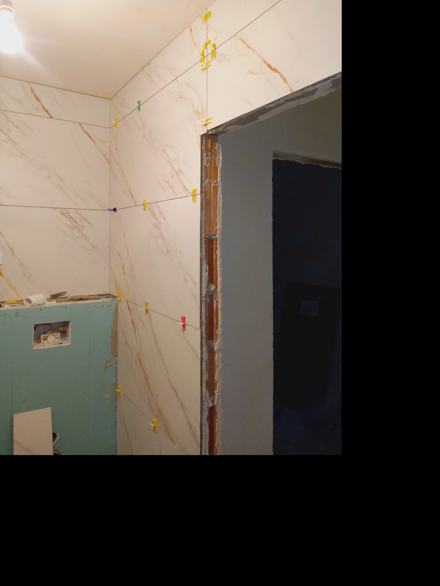 Proces układania płytek imitujących marmur w łazience, z użyciem poziomicy i krzyżyków dystansowych. Widoczne elementy instalacji podtynkowej.