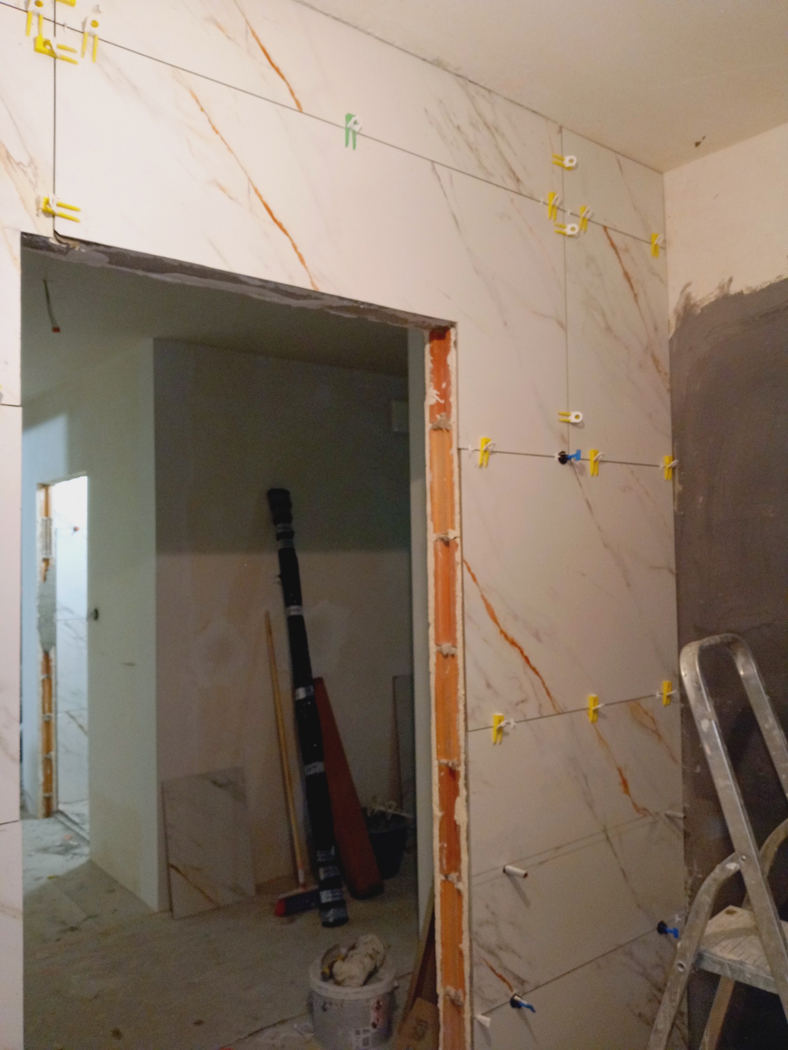 Układanie płytek imitujących marmur na ścianie, z widocznymi krzyżykami dystansowymi i poziomowaniem. W tle widoczne przygotowania do dalszych prac wykończeniowych.