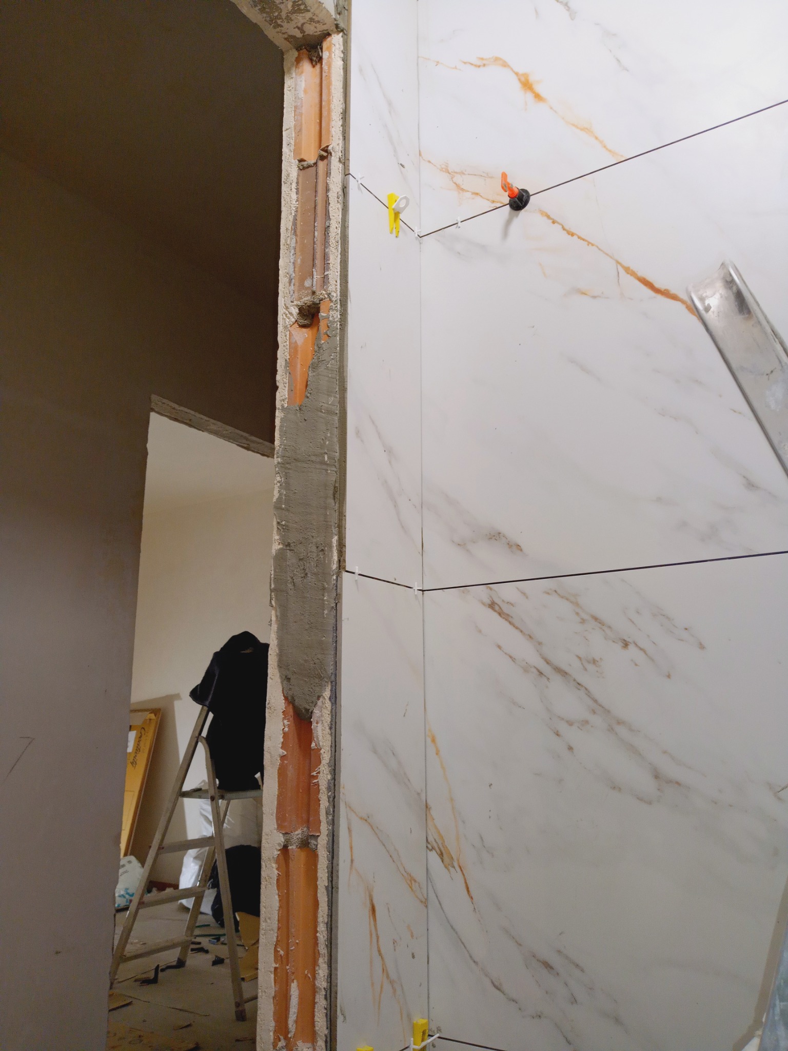 Ściana z płytek imitujących marmur w trakcie montażu, z użyciem poziomicy i klipsów poziomujących. Widoczny fragment ościeżnicy drzwiowej w surowym stanie.