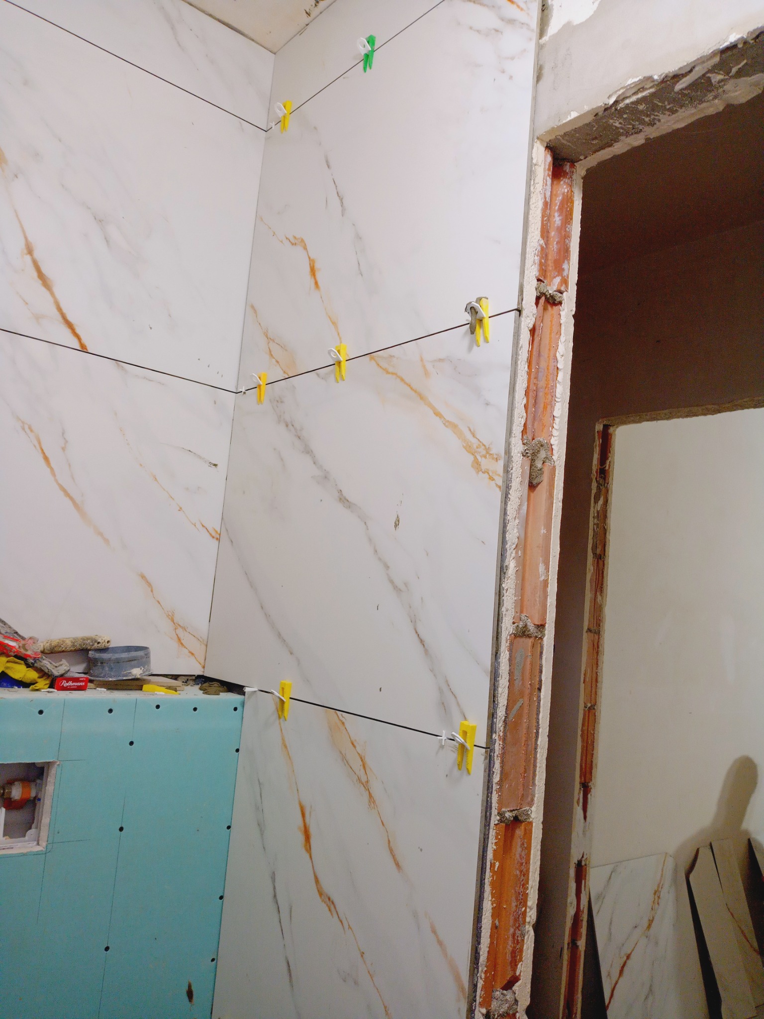 Montaż płytek imitujących marmur w łazience, z użyciem klipsów poziomujących. Widoczna płyta kartonowo-gipsowa i surowa ościeżnica drzwi.