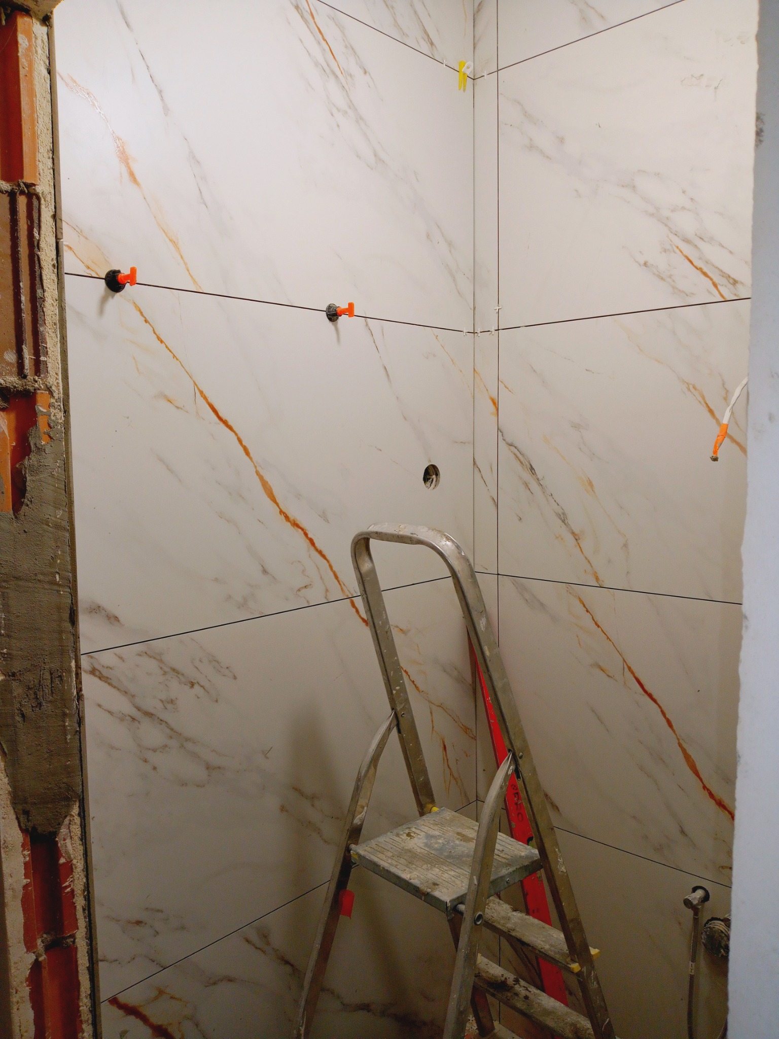 Układanie płytek imitujących marmur w rogu łazienki. Widoczna drabina, system poziomowania płytek i fragment ściany z instalacją.