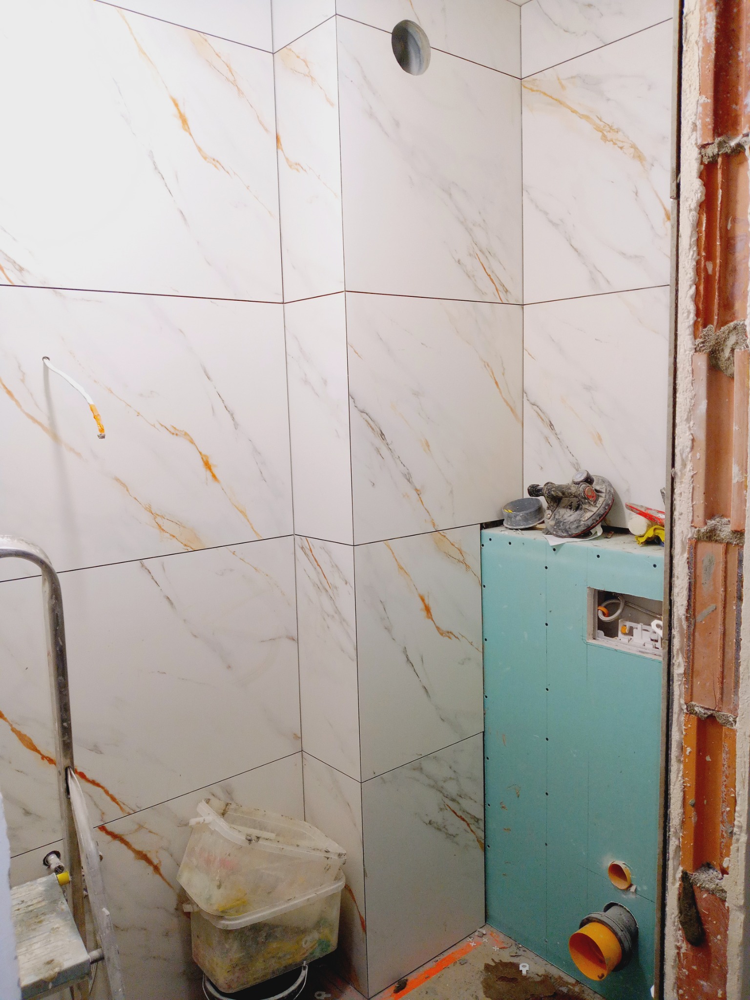 Białe płytki imitujące marmur z pomarańczowymi żyłkami w łazience, częściowo wykończone. Widoczne narzędzia i płyta karton gips na ścianie.