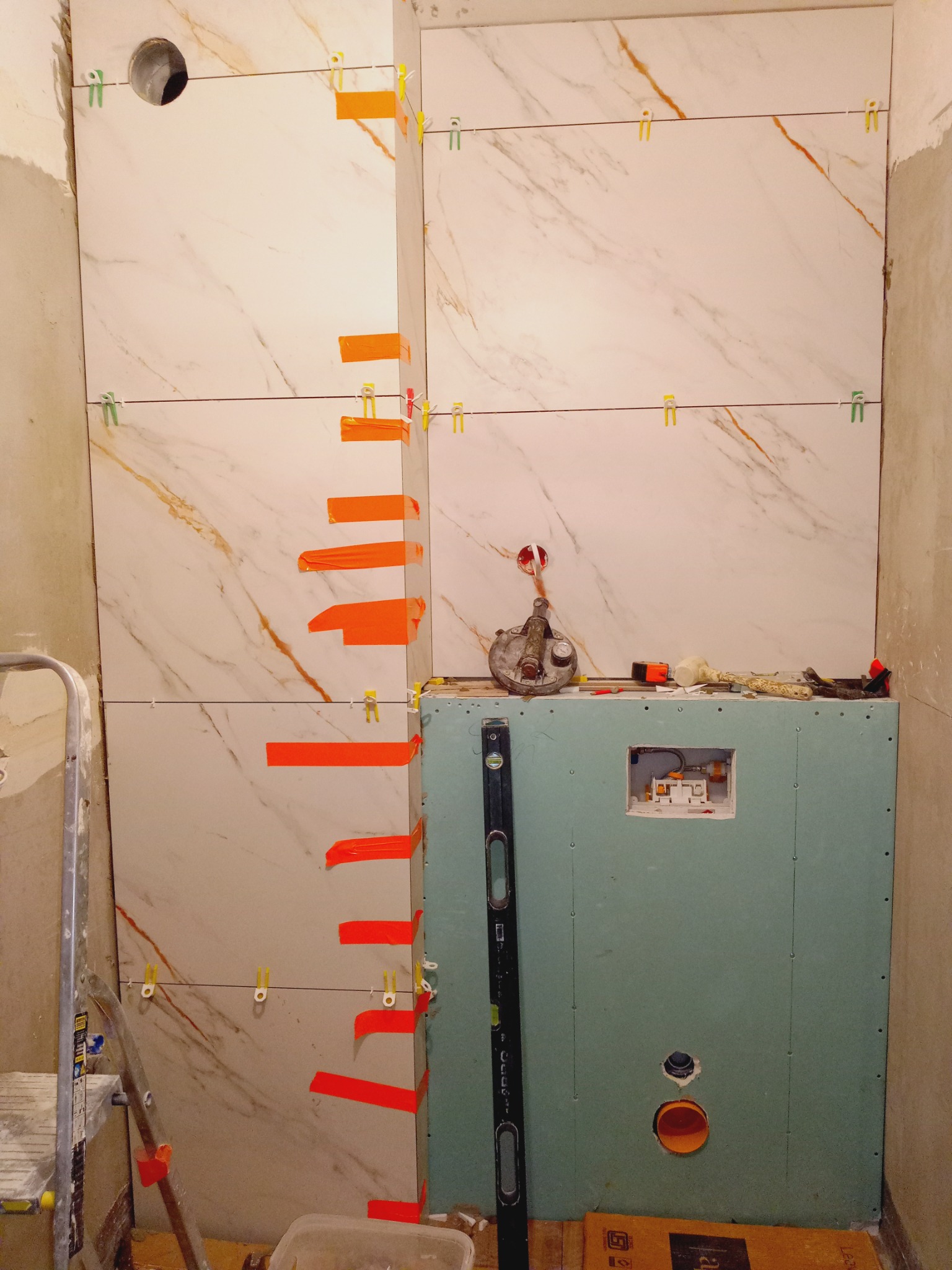 Ściana z marmurowych płytek w trakcie montażu, zabezpieczona pomarańczową taśmą i klipsami, z widoczną instalacją hydrauliczną i narzędziami glazurniczymi.