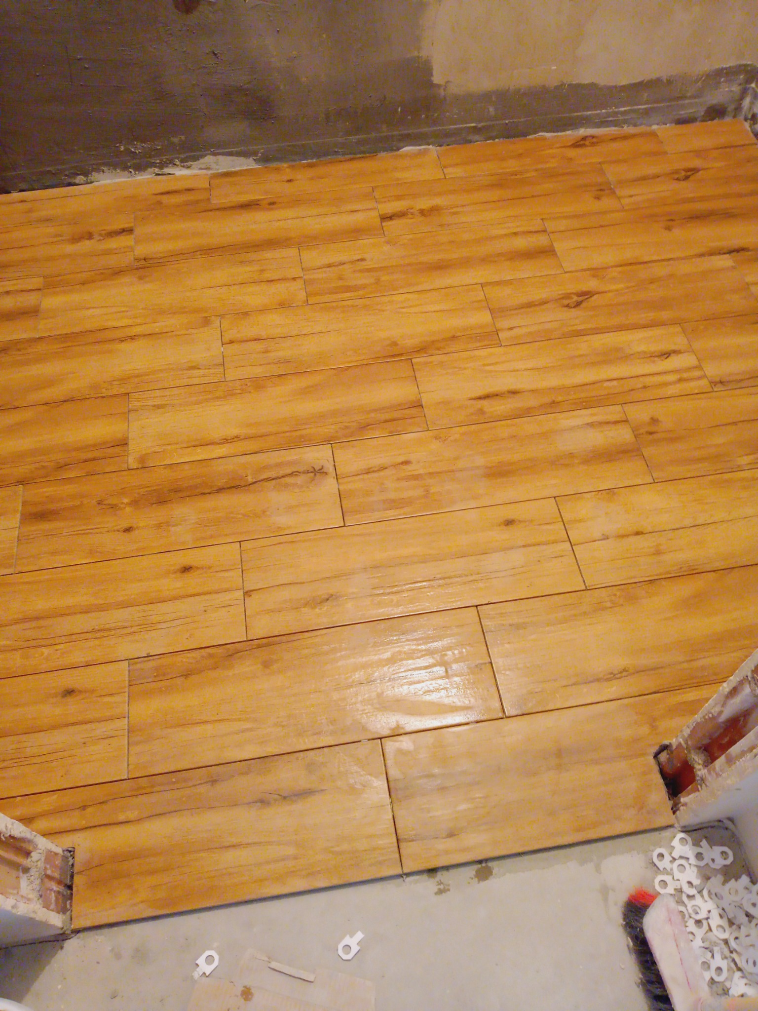 Ułożone, podłużne płytki imitujące drewno na podłodze w trakcie remontu. Widoczne krzyżyki dystansowe i fragment ściany w surowym stanie. Ciepła kolorystyka płytek.