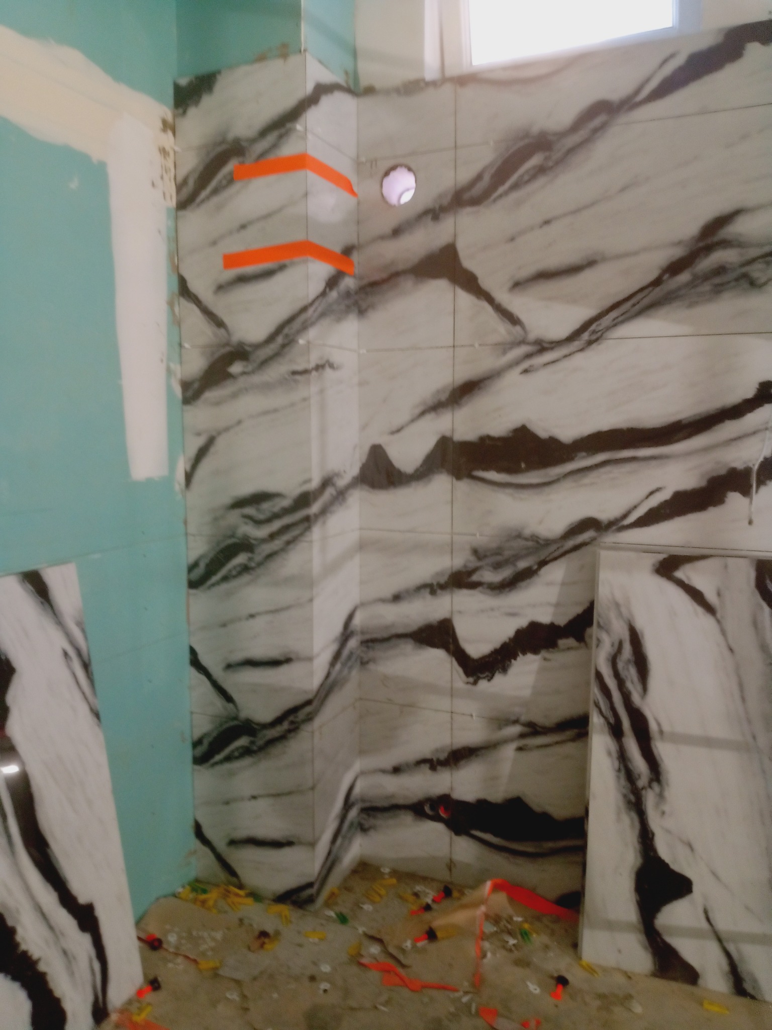 Montaż płytek imitujących marmur na ścianie w łazience, widoczne pomarańczowe kliny dystansowe i fragment płyty karton-gips. Prace glazurnicze w toku.