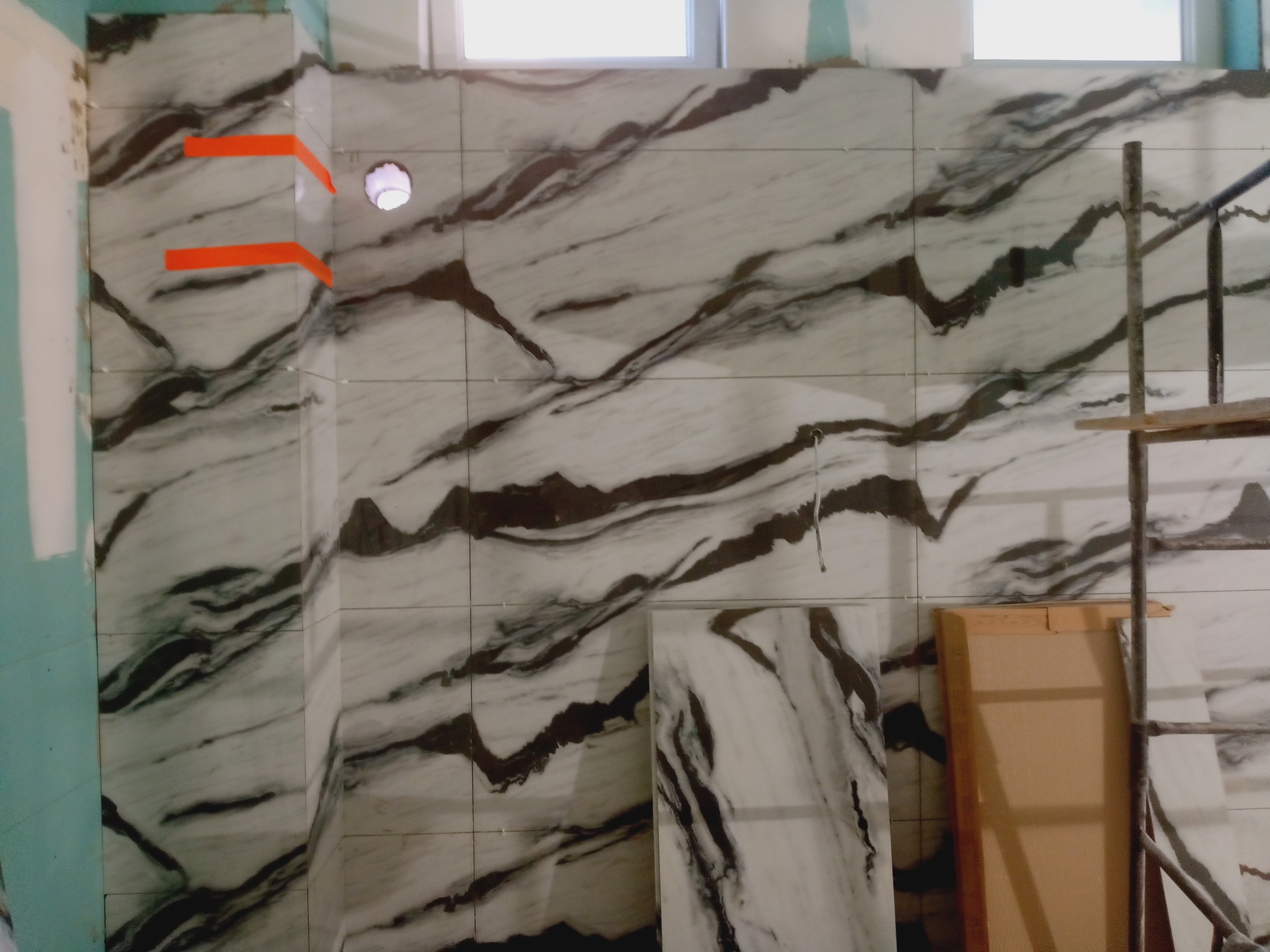 Ściana łazienki wyłożona białymi płytkami z szarym marmurkowym wzorem. Widoczne pomarańczowe kliny dystansowe i otwór na instalację. Fragment rusztowania po prawej stronie.