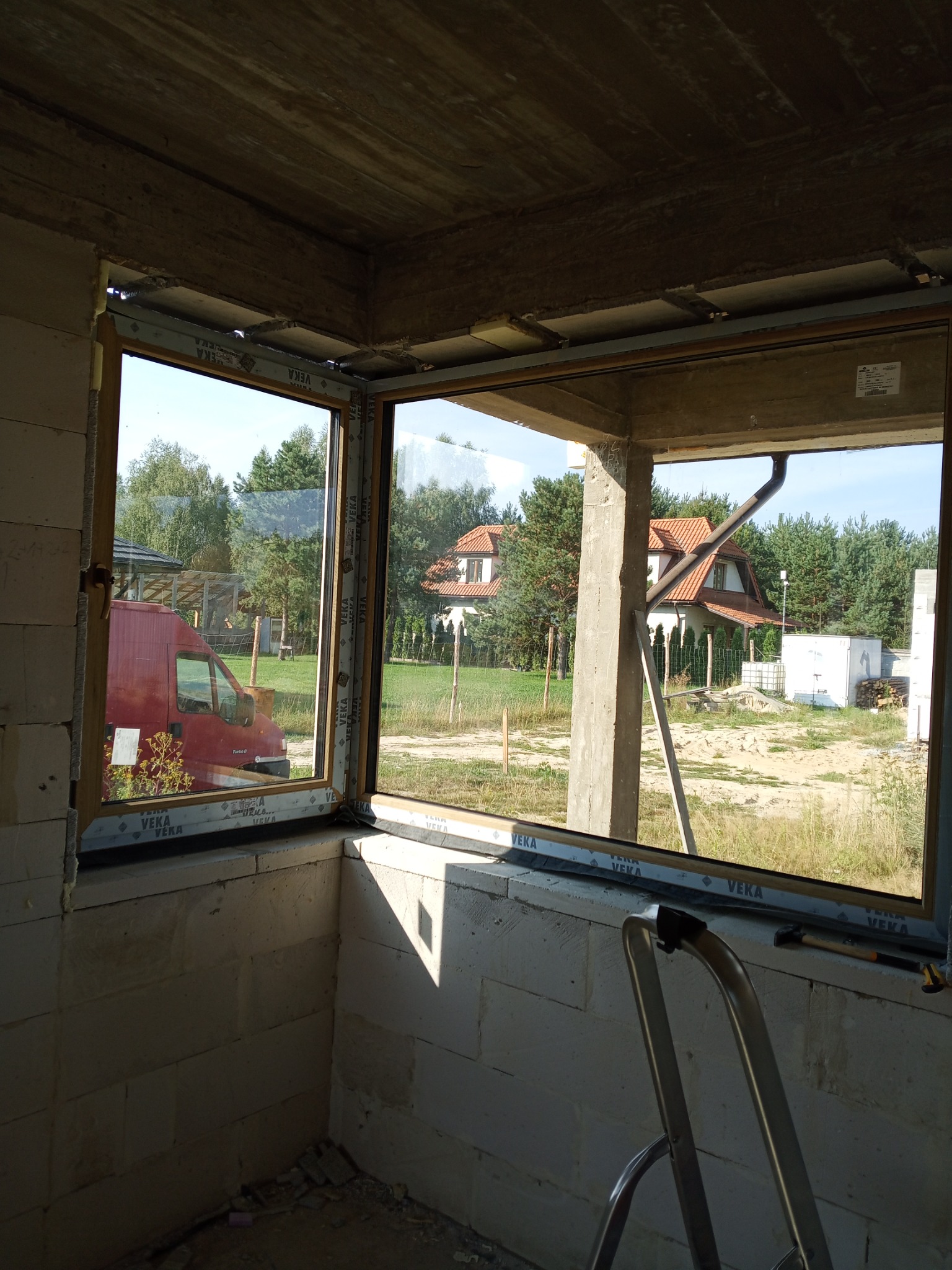 Nowe okno PCV z widokiem na dom jednorodzinny i zieleń, zamontowane w surowym wnętrzu z betonu komórkowego. Widoczna drabina i narzędzia.