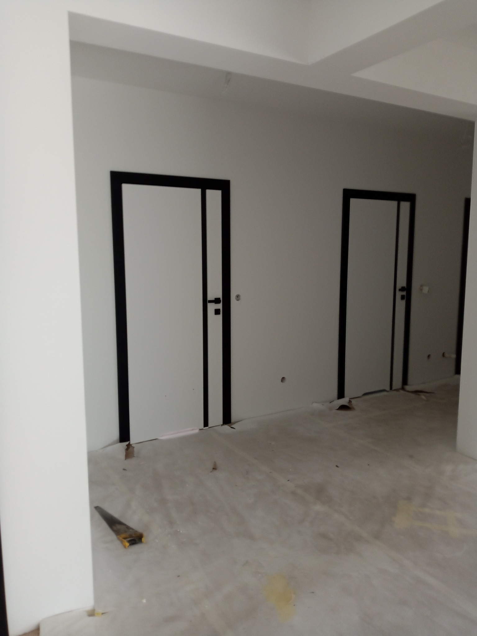Wnętrze w trakcie wykańczania: białe ściany, dwoje białych drzwi z czarnymi ościeżnicami. Na podłodze leży piła ręczna. Minimalistyczny design.