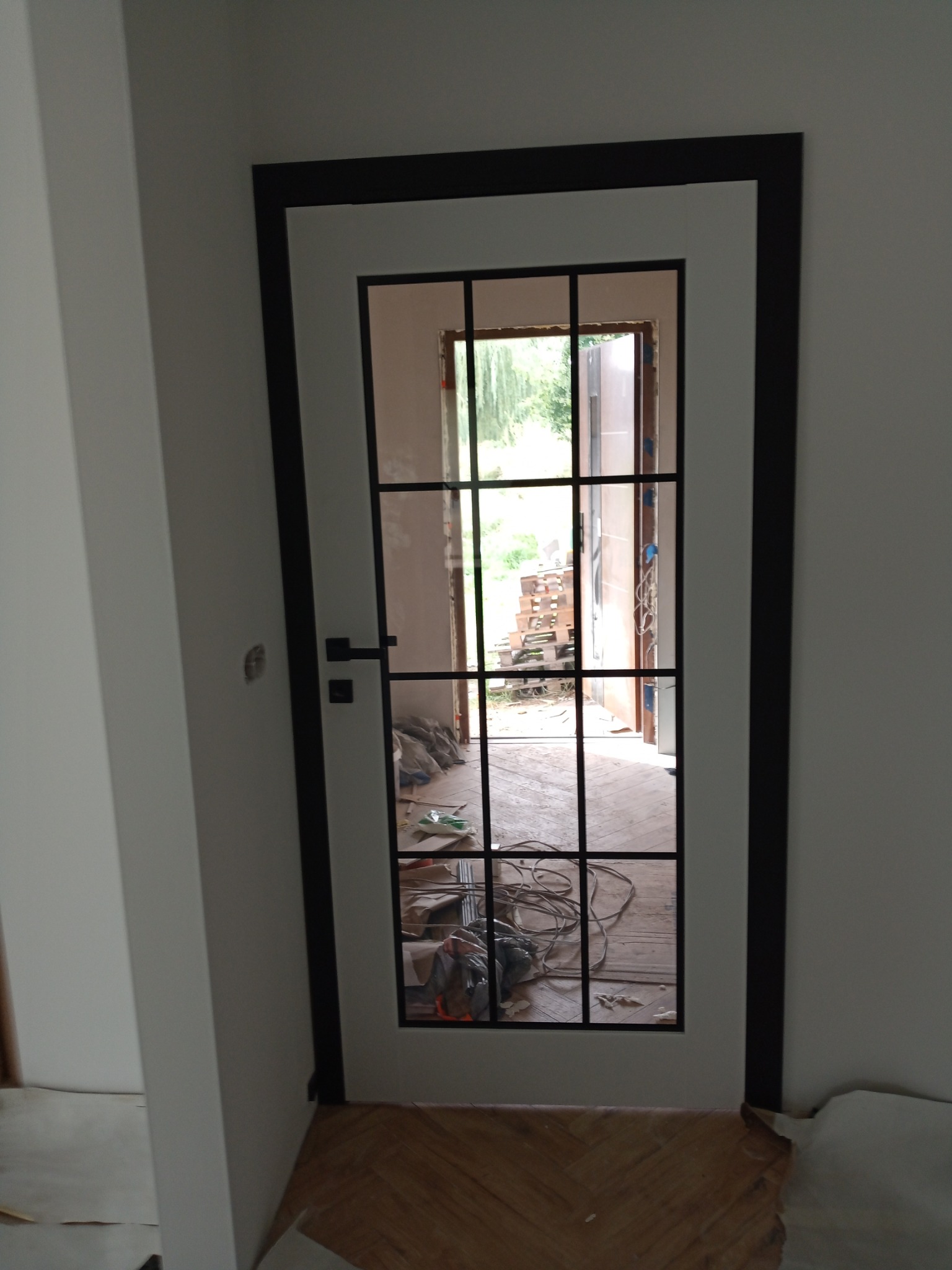 Nowoczesne, białe drzwi z czarną ramą i szprosami, widok na zewnątrz przez szklane panele. Podłoga częściowo zabezpieczona, widoczne kable i narzędzia.