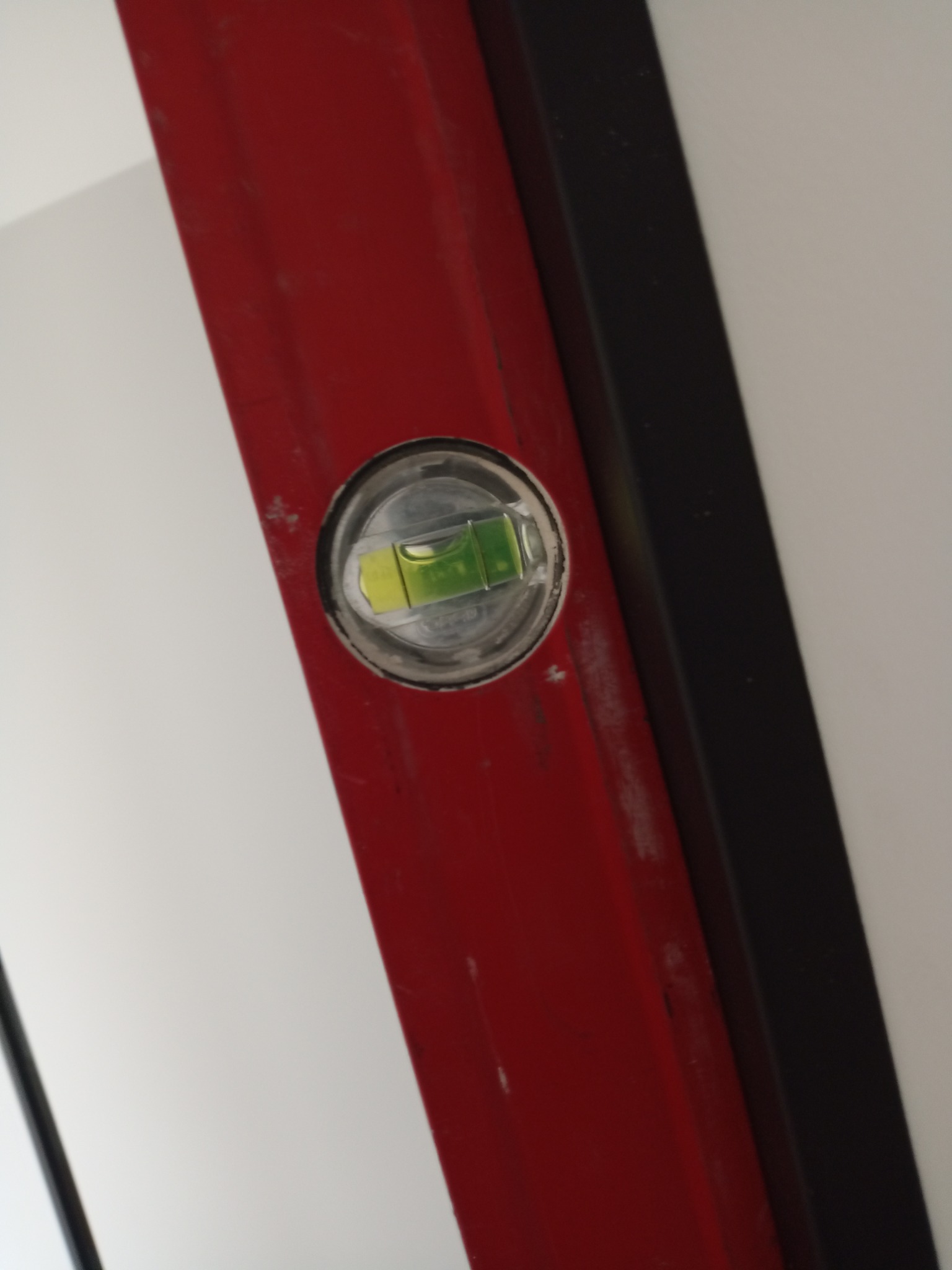 Czerwona poziomica przyłożona do ściany z czarną listwą, wskazująca wypoziomowanie. Detal narzędzia pracy podczas wykańczania wnętrz.