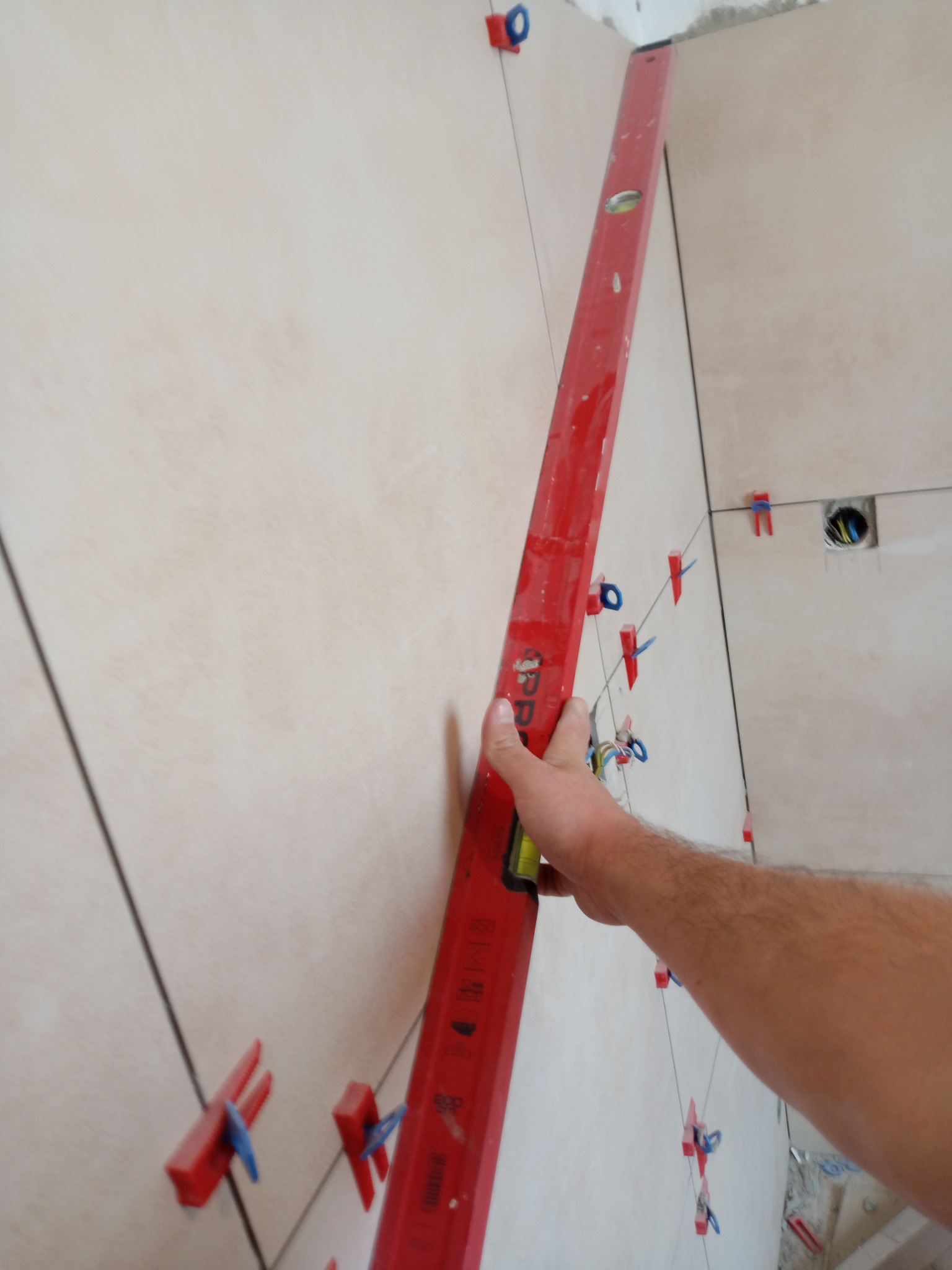 Układanie jasnych płytek na ścianie z użyciem poziomicy i klipsów dystansowych. Widoczny fragment dłoni przytrzymującej narzędzie.