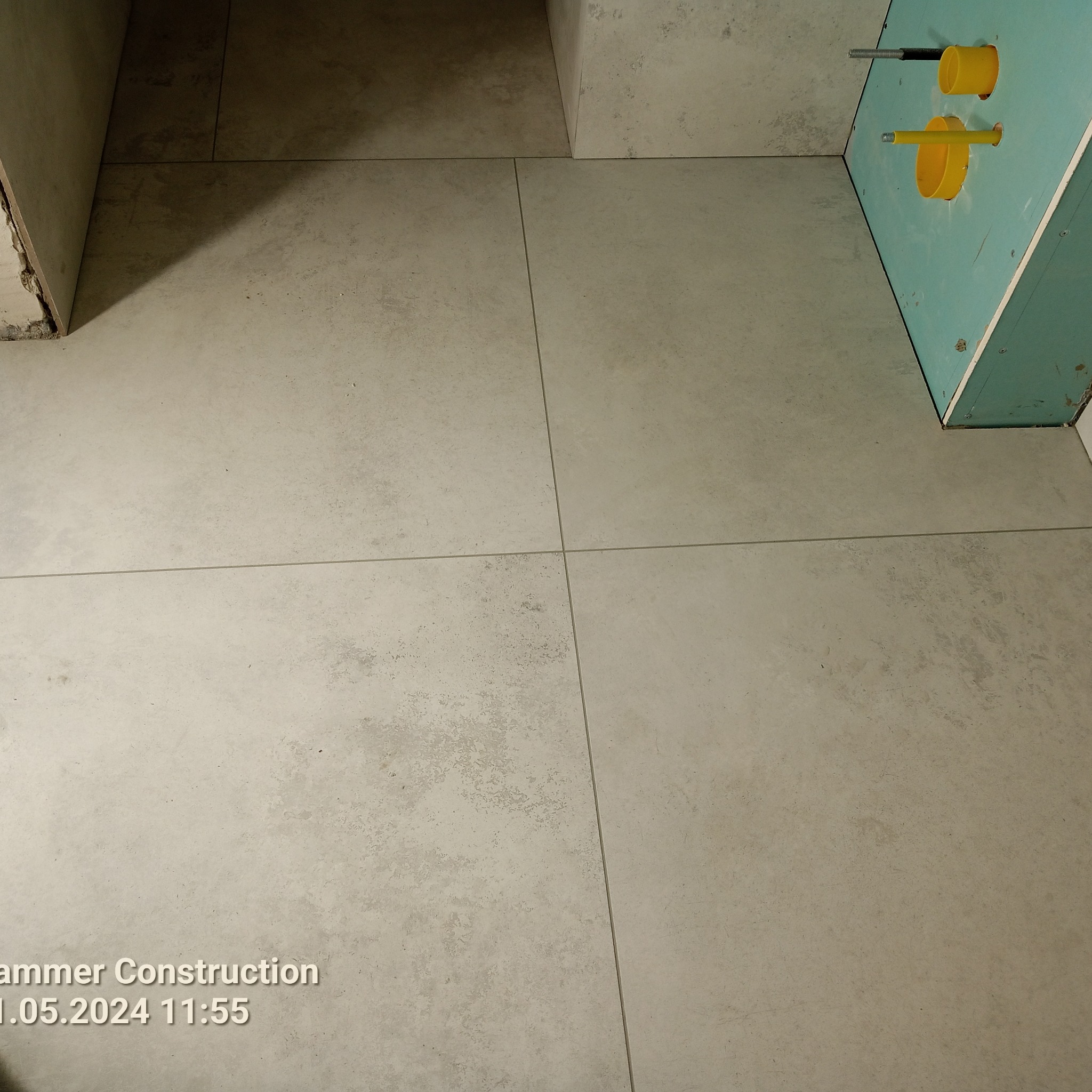 Ułożone, beżowe płytki podłogowe w łazience, widoczne fragmenty ściany z instalacją wodną. Minimalistyczny design, neutralna kolorystyka.