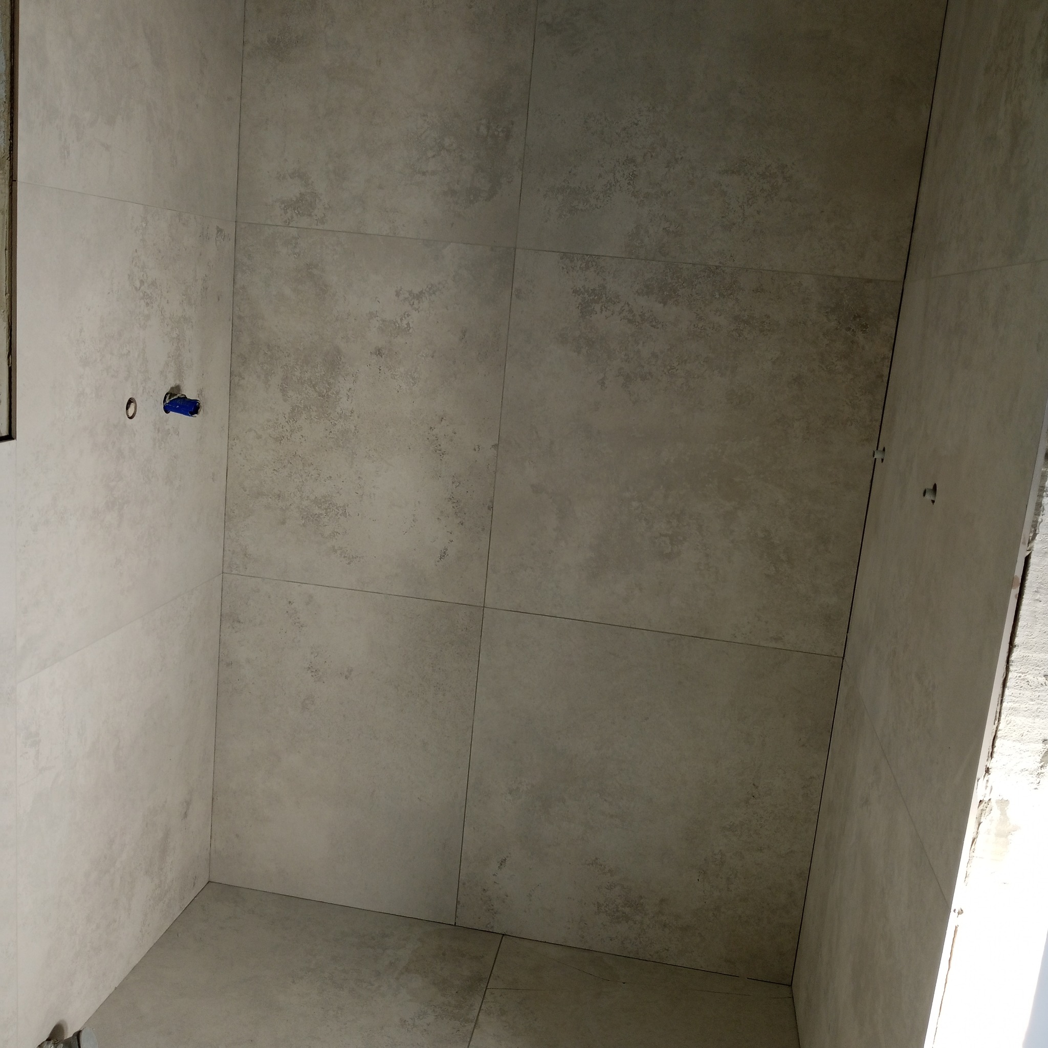Wykończona ściana i podłoga w łazience z szarymi, matowymi płytkami imitującymi beton. Widoczne przyłącze wodne i fragment surowej ściany.