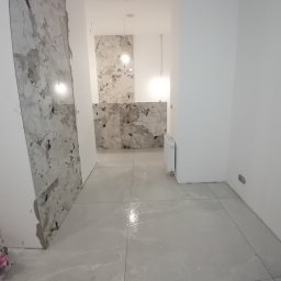 DOM-ART - Wnętrze w trakcie prac: szare płytki podłogowe i marmurowe na ścianie, widoczny grzejnik. Częściowo wykończona ściana z niedociągnięciami przy podłodze.