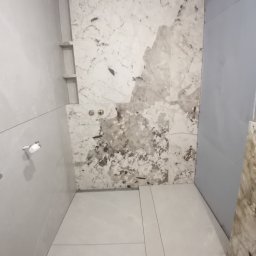 DOM-ART - Wnętrze łazienki w trakcie prac wykończeniowych. Widoczne płytki imitujące marmur na ścianach i podłodze, oraz przygotowane miejsce na półki wnękowe.