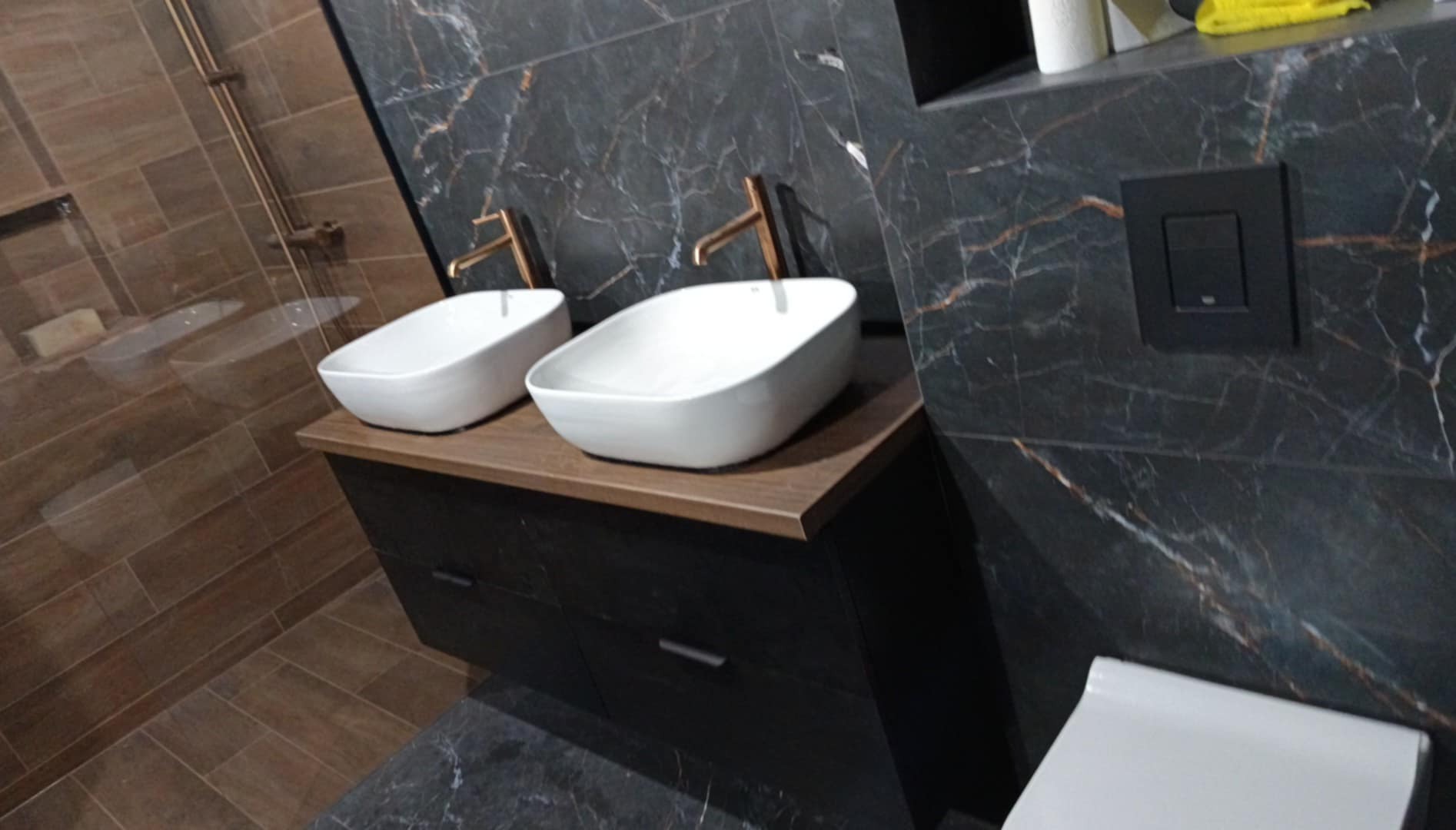 Nowoczesna łazienka z dwoma białymi umywalkami nablatowymi i złotą armaturą na tle płytek imitujących marmur. Ciemna szafka pod umywalkami i brązowe płytki na ścianie prysznica.