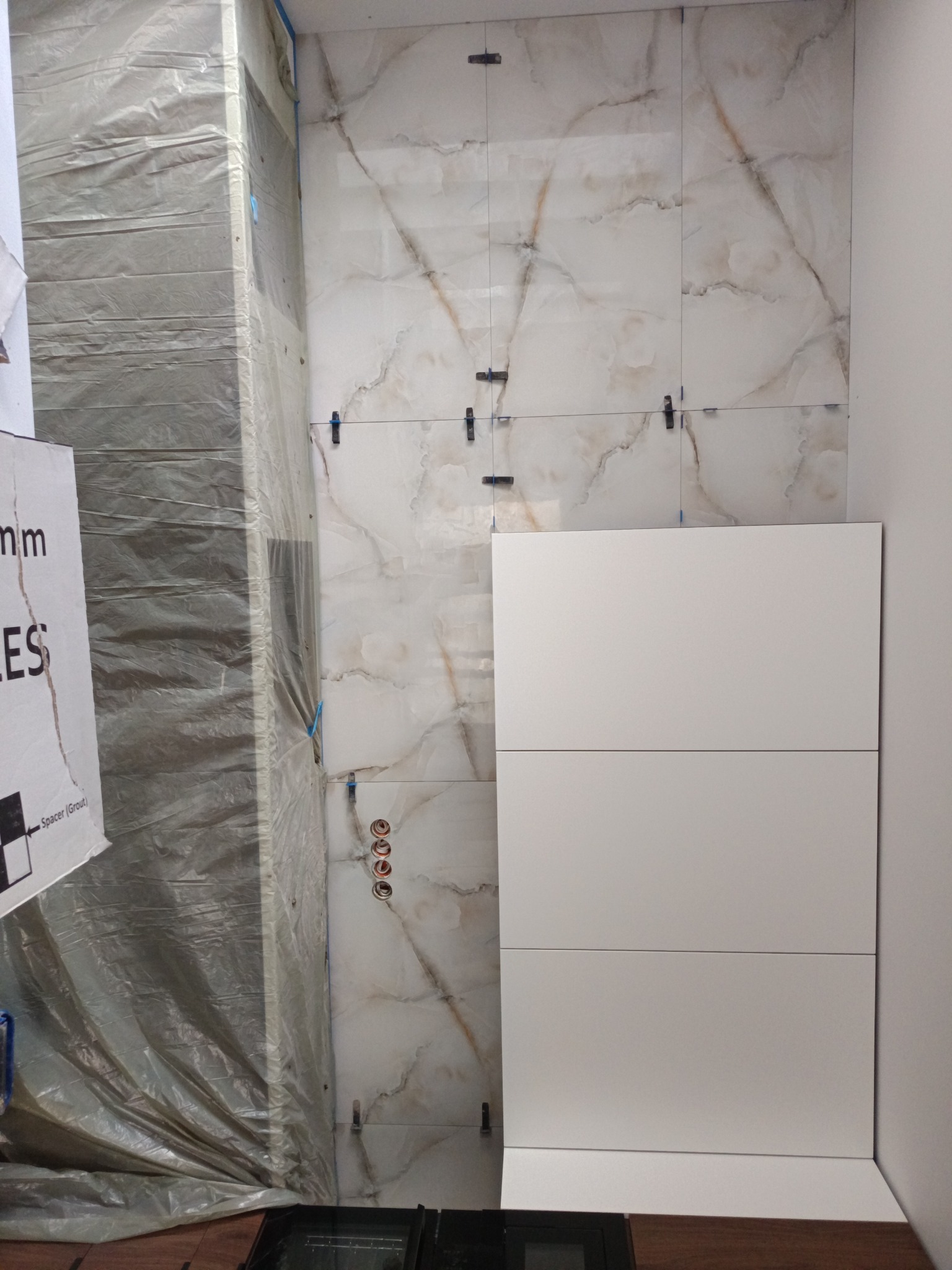 Ściana łazienki z marmurowymi płytkami w trakcie montażu, widoczne krzyżyki dystansowe i zabezpieczona folią kolumna, obok biała zabudowa meblowa.