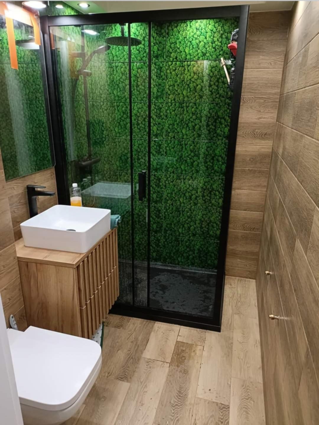 Nowoczesna łazienka z kabiną prysznicową typu walk-in, wykończona płytkami imitującymi drewno i ścianą z mchu. Umywalka nablatowa na drewnianej szafce.