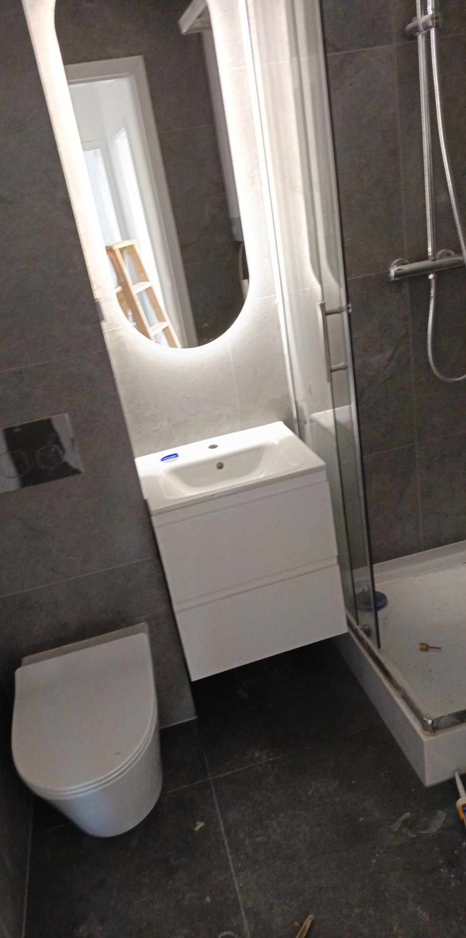 Wykończona łazienka z szarą płytką, białą umywalką z szafką i toaletą. W tle podświetlane lustro i kabina prysznicowa. Widoczne detale wykończeniowe.