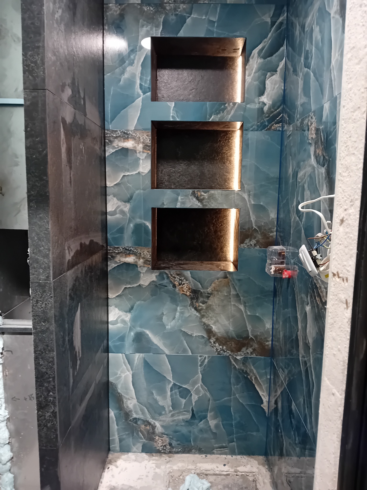Błękitna ściana z płytek imitujących marmur, z trzema wnękami podświetlonymi ciepłym światłem. Widoczne przewody elektryczne i fragment ściany w trakcie prac wykończeniowych.