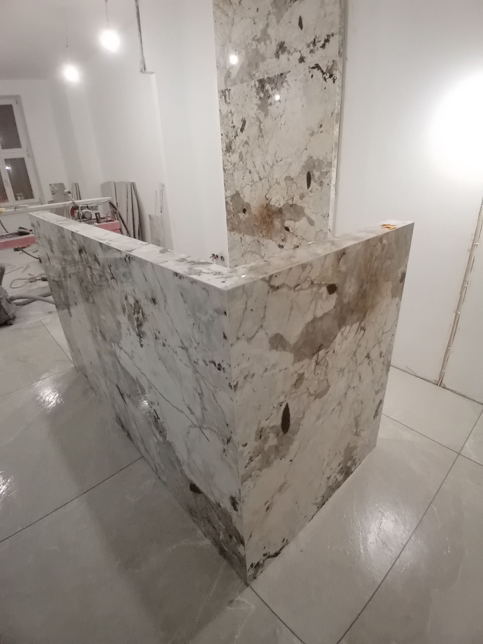 Wykończona ściana i podłoga z płytek imitujących marmur w jasnej tonacji. Widoczne narzędzia glazurnicze w tle. Nowoczesny design.