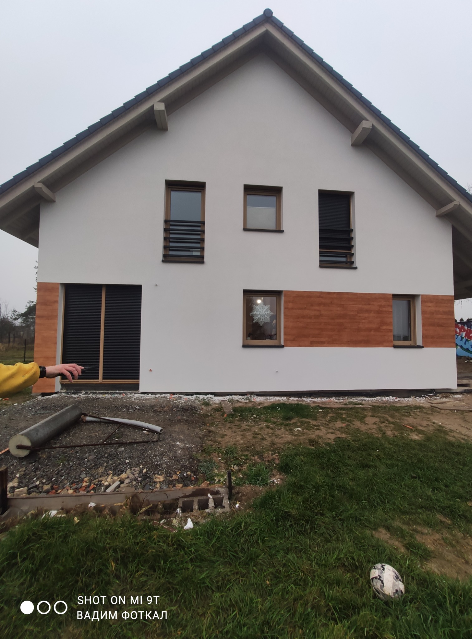 Nowoczesny dom z białą elewacją i drewnianymi akcentami. Widoczne okna z roletami i dekoracja okienna. Na pierwszym planie fragment terenu z elementami budowlanymi.