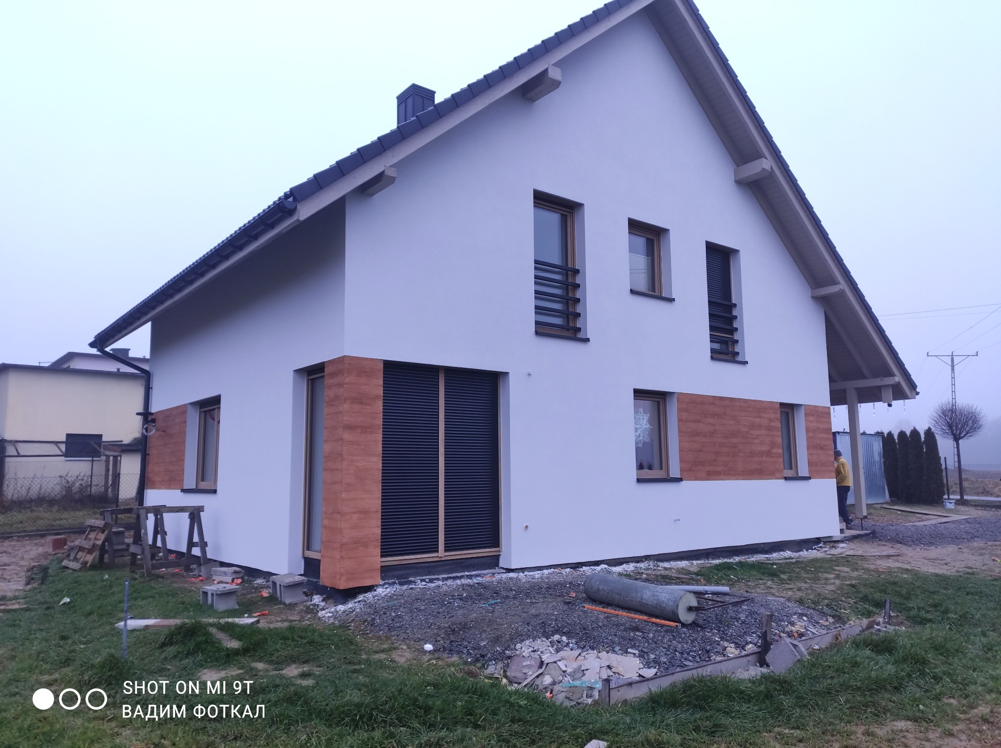 Nowoczesny dom jednorodzinny z białą elewacją i elementami dekoracyjnymi z paneli imitujących drewno. Widoczne rusztowania i materiały budowlane sugerują trwające prace wykończeniowe.