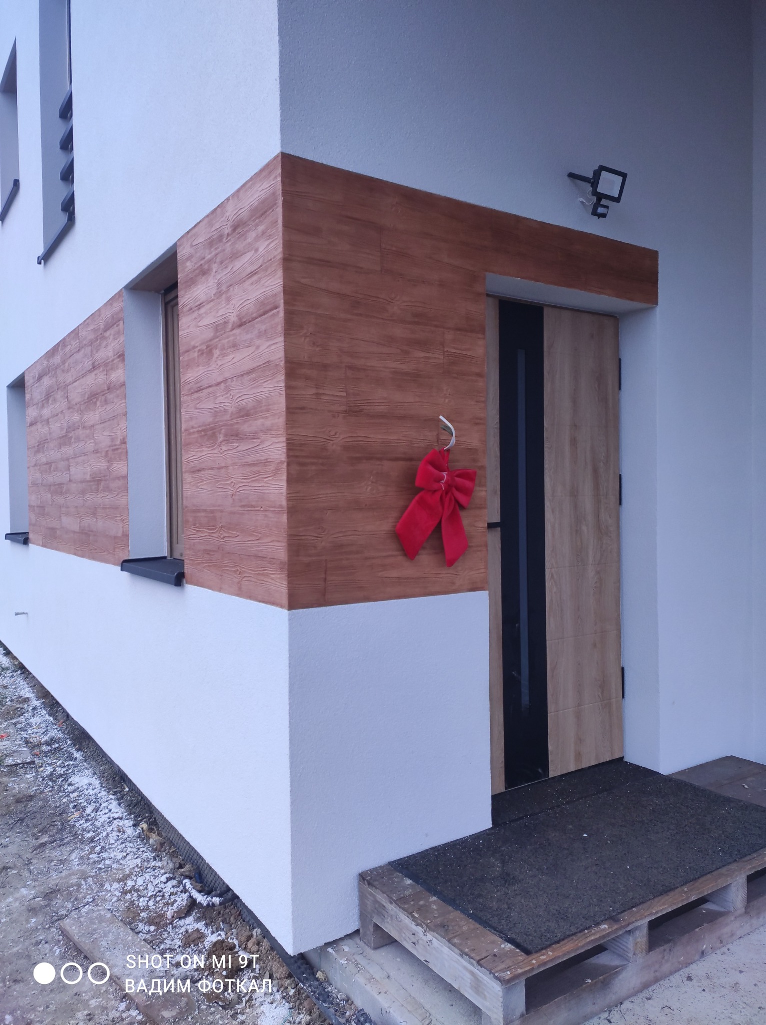 Nowoczesny dom z elewacją imitującą drewno, ozdobiony czerwoną kokardą przy drzwiach wejściowych. Widoczny fragment okna i oświetlenie zewnętrzne.