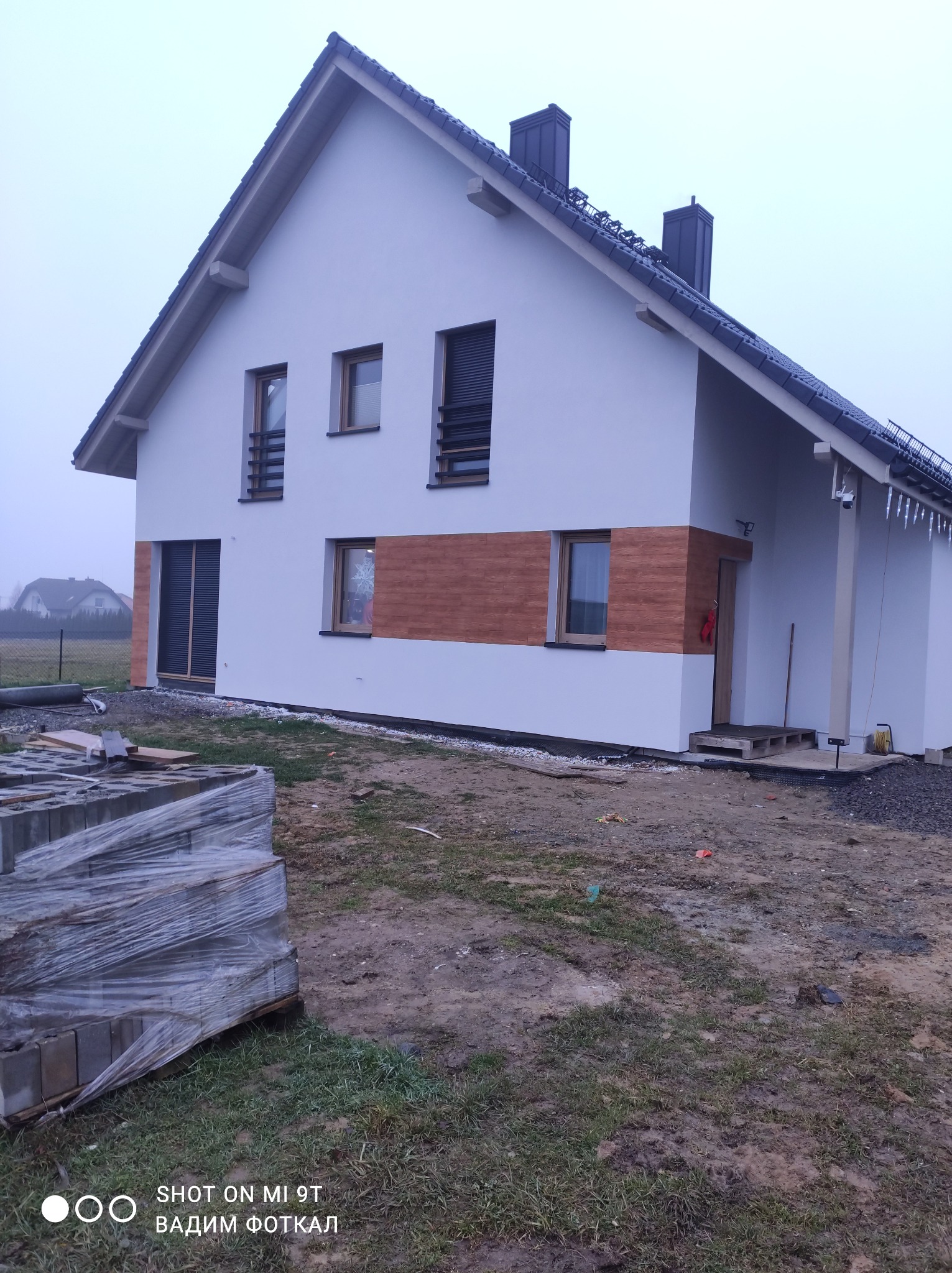 Nowoczesny dom jednorodzinny z białą elewacją i drewnianymi akcentami. Widoczne okna z roletami i kostka brukowa na pierwszym planie. Dom w trakcie wykańczania.