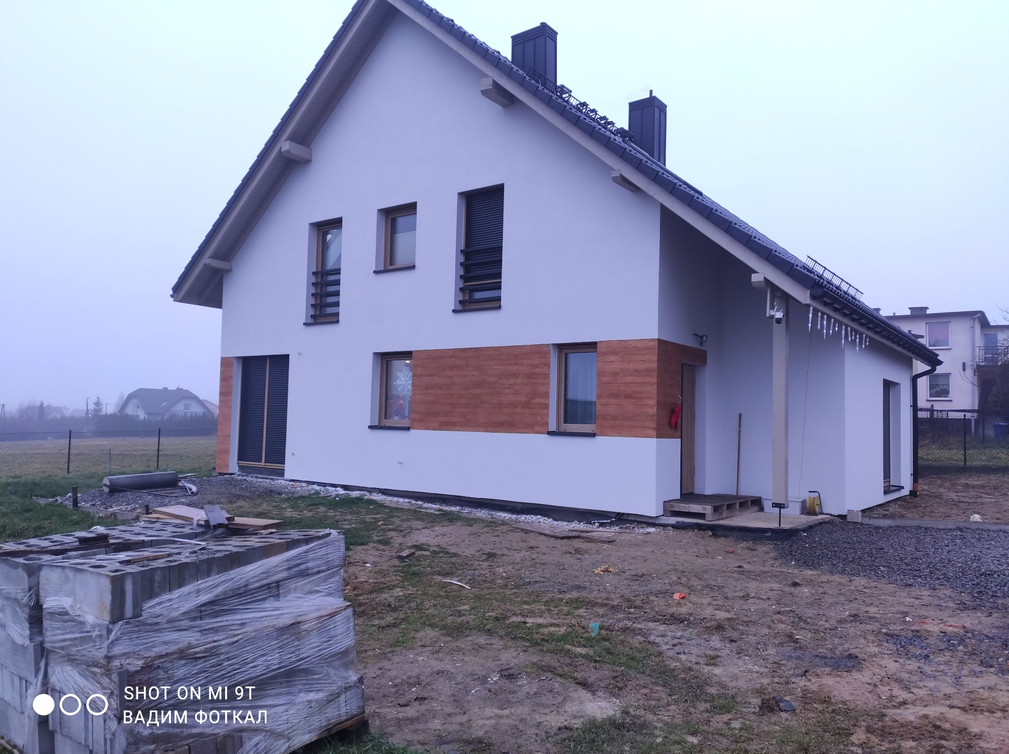 Nowoczesny dom jednorodzinny z białą elewacją i drewnianymi akcentami, widoczny stos bloczków budowlanych na pierwszym planie, pochmurny dzień.