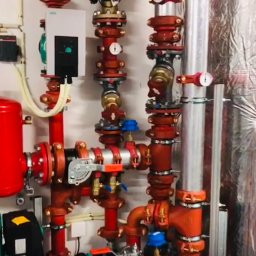 USŁUGI HYDRAULICZNE Filip Turo - Złożony system rur i zaworów w kolorze czerwonym i srebrnym na białej ścianie, z elementami izolacji. Widoczne manometry i urządzenia sterujące.