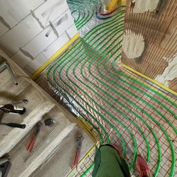 USŁUGI HYDRAULICZNE Filip Turo - Zielone rury ogrzewania podłogowego ułożone na folii, widoczne narzędzia hydrauliczne i fragmenty ścian w surowym stanie. Żółta taśma na obrzeżach.